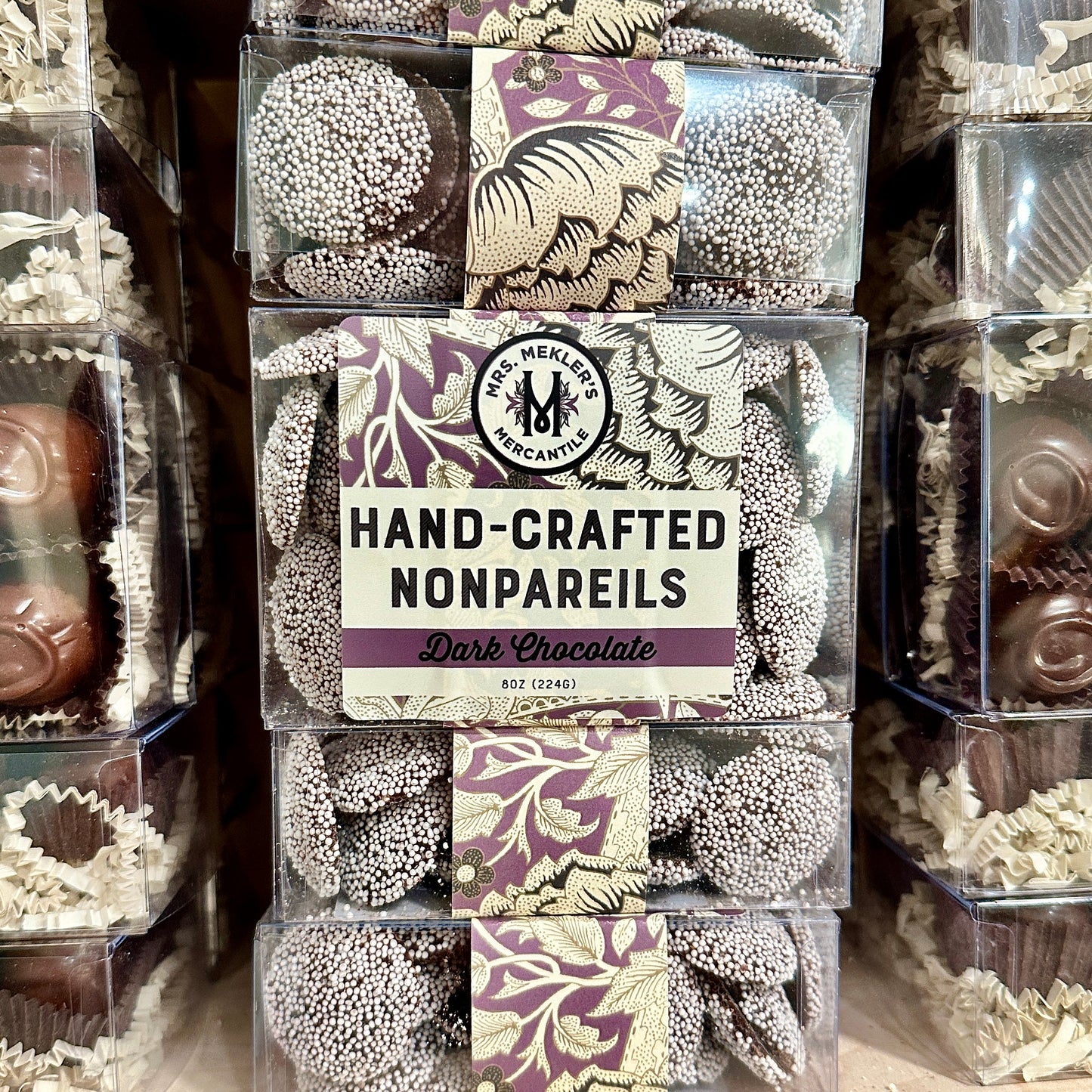 A clear plastic box containing dark chocolate nonpareils with visible white nonpareil sprinkles on top. The packaging features a label with the brand name and the product description.