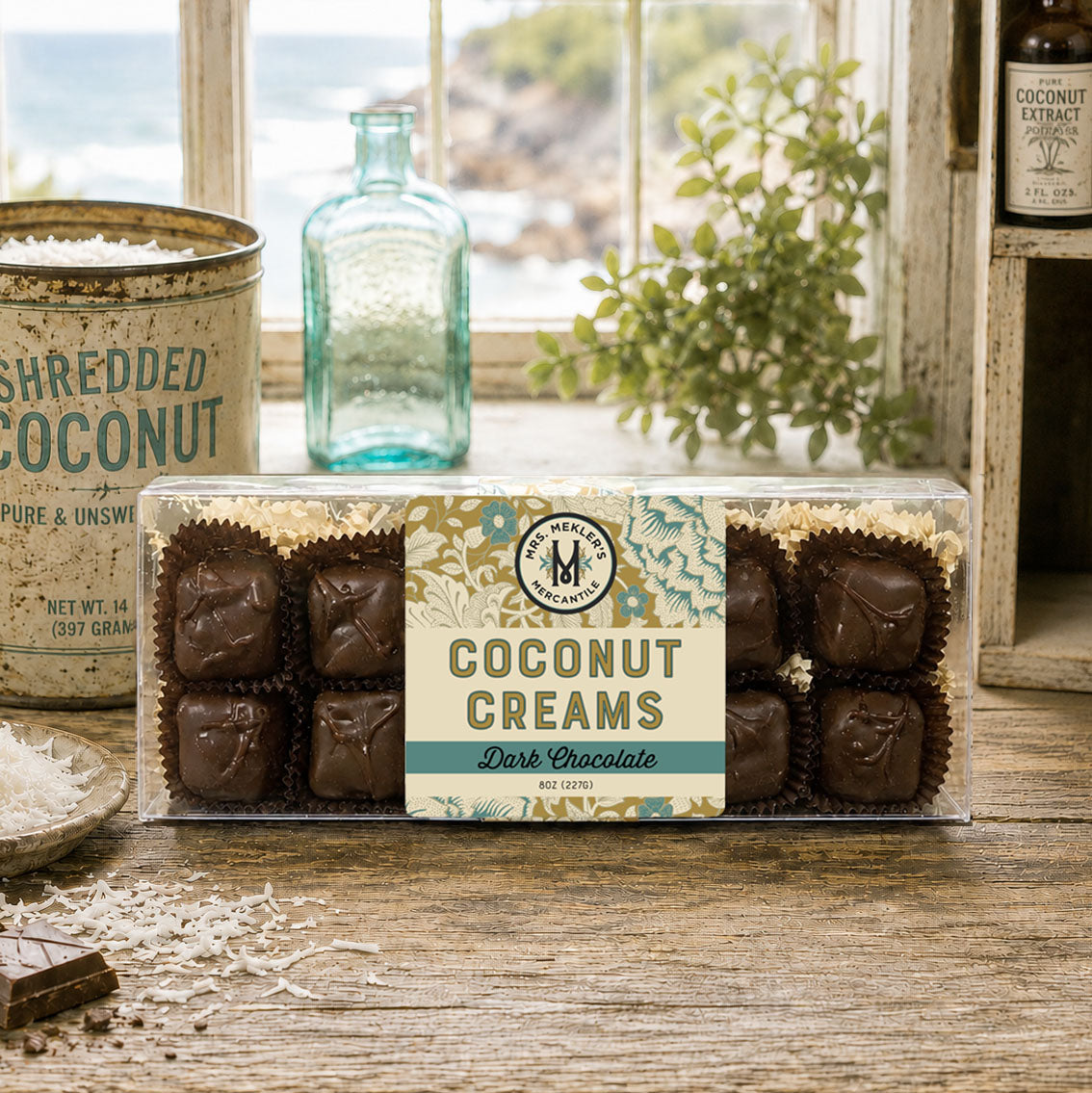 A clear plastic box of Mrs Mekler's Mercantile Coconut Creams Dark Chocolate sits on a rustic wooden table with a vintage tin, a small plate of shredded coconut, and a blue glass bottle in the background near a window.