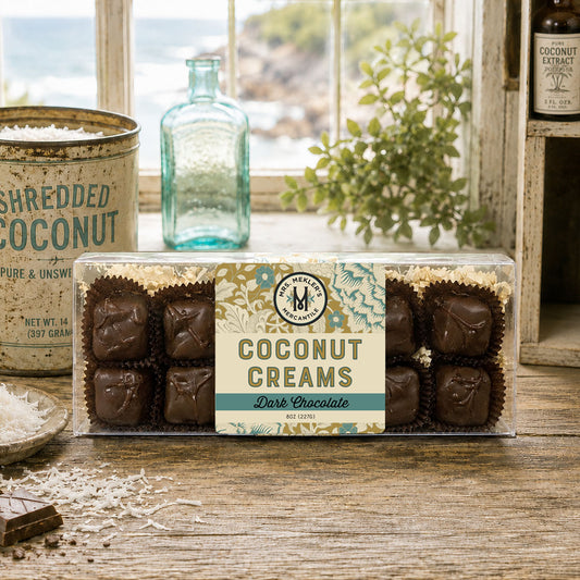 A clear plastic box of Mrs Mekler's Mercantile Coconut Creams Dark Chocolate sits on a rustic wooden table with a vintage tin, a small plate of shredded coconut, and a blue glass bottle in the background near a window.