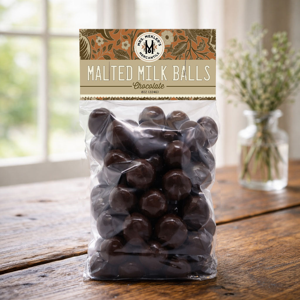 A clear plastic bag of chocolate malted milk balls sits on a rustic wooden table, with a blurred window and a small vase of flowers in the background. The packaging features a decorative label at the top that reads 'Malted Milk Balls Chocolate' and shows the 'Malted Milk Company' logo.