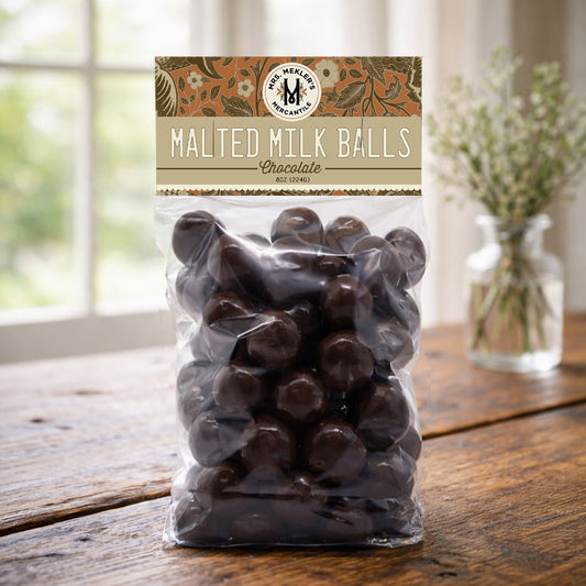 A clear plastic bag of chocolate malted milk balls sits on a rustic wooden table, with a blurred window and a small vase of flowers in the background. The packaging features a decorative label at the top that reads 'Malted Milk Balls Chocolate' and shows the 'Malted Milk Company' logo.