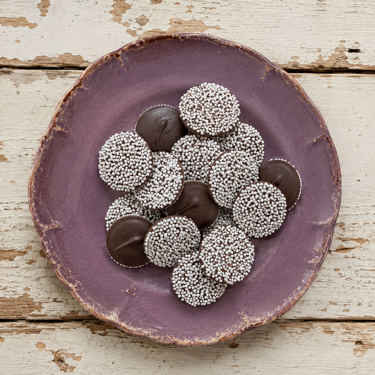 A top-down view of dark chocolate nonpareils presented on a rustic, textured purple ceramic plate over a distressed white wooden background.