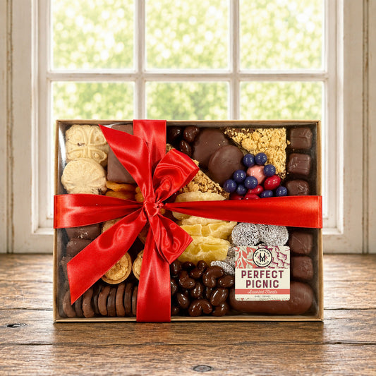 A gourmet chocolate and cookie gift box filled with assorted treats, decorated with a large red satin bow, and labeled 'Perfect Picnic'. The box is set on a rustic wooden table in front of a bright window.