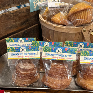 Several individually wrapped cinnamon icelandic muffins with colorful labels are displayed on a metal tray, with a wooden basket of muffins in the background.