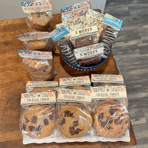 A bakery assortment of packaged cookies and s'mores arranged on a wooden table. The set includes Heath Bag Crunch Cookies, Colossal Cookies, and chocolate-dipped s'mores in clear bags and a blue bowl.