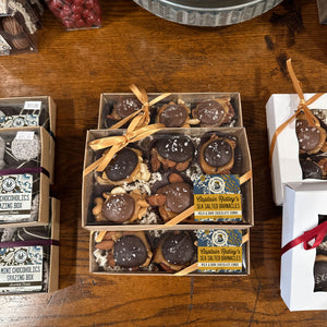 Three open boxes of Captain Ridley's Sea Salted Barnacles arranged on a wooden table. The center box shows round chocolate-covered barnacle with almonds, white sea salt flakes, and a gold ribbon.