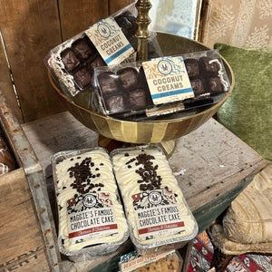 Two packaged loaves of Maggie's Famous Chocolate Cake are displayed on a rustic wooden table next to a gold pedestal bowl containing chocolate truffles and packaged coconut creams. The setting features a vintage wooden chest and a green cushion in the background.