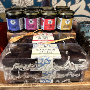 A gift assortment display featuring jars of preserves and boxes of chocolate patties. In the back, six glass jars with gold lids are labeled Wild Blueberry Chutney, Tart & Tangy Chutney, Cherry Amaretto Chutney, and Butter Rum Brilliance