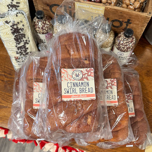 Several packages of cinnamon swirl bread arranged on a wooden table, with a larger loaf in the center and smaller loaves around it. The bread is wrapped in clear plastic with visible labels reading 'Cinnamon Swirl Bread' and 'Small Batch.